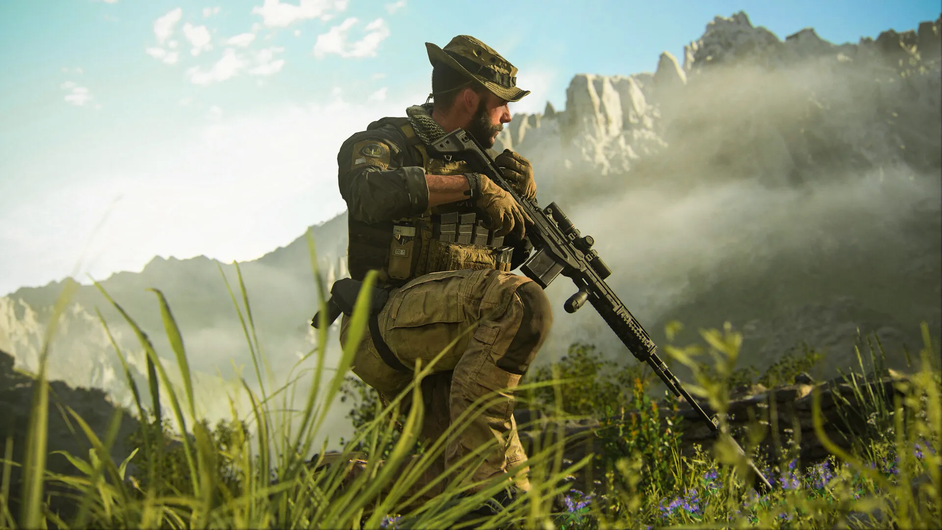 A bearded soldier in a boonie hat kneels in tall grass with a scoped rifle, mountains behind